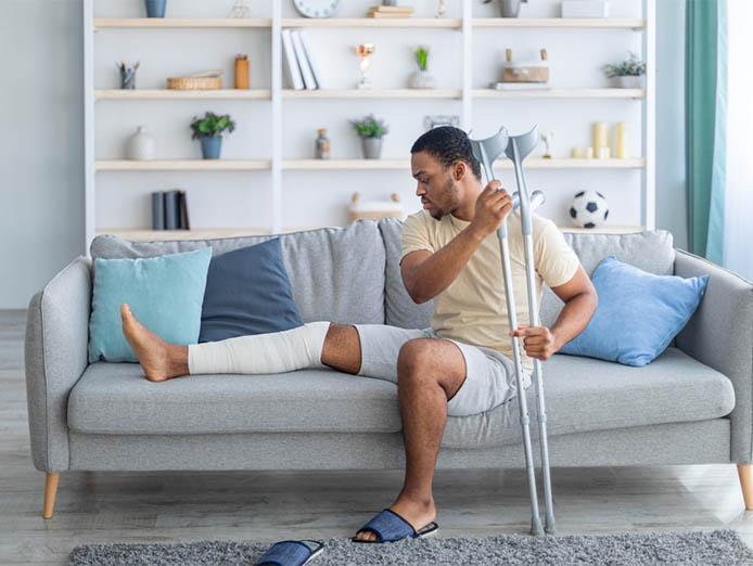 oung African American guy with plastered leg sitting on sofa, leaning on crutches at home